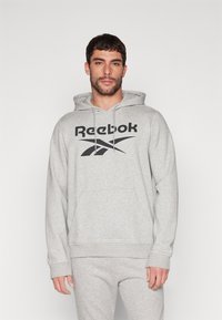 Pilkas džemperis su priekine kišene, kapuche su raišteliais ir juodu Reebok logotipu. Audinys atrodo minkštas su šviesia heather tekstūra.