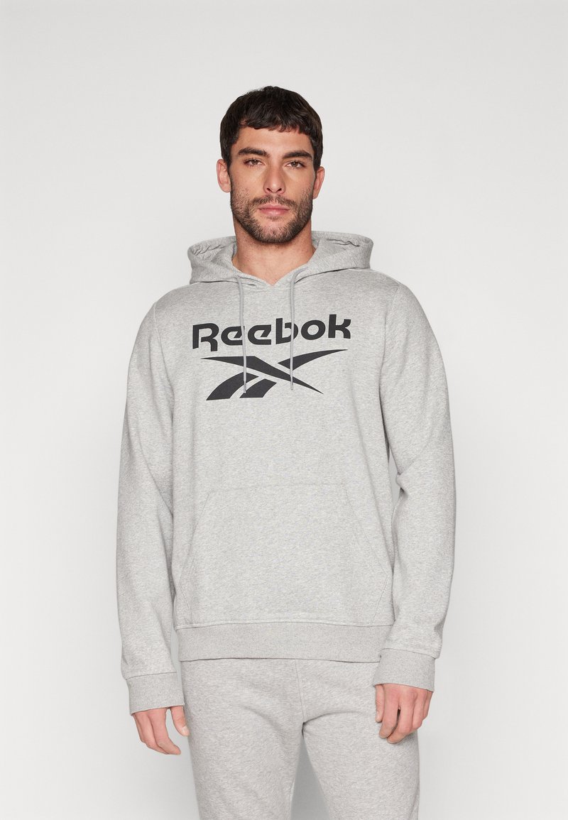 Pilkas džemperis su priekine kišene, kapuche su raišteliais ir juodu Reebok logotipu. Audinys atrodo minkštas su šviesia heather tekstūra.