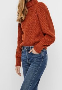Pull col roulé en maille orange brûlé avec un motif texturé diagonal. Associé à un jean bleu taille haute avec poches.