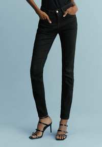 Femme portant un jean skinny noir taille haute et des sandales noires à talons hauts avec des brides, debout les mains dans les poches devant un fond bleu pâle.