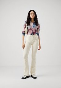 Långärmad tröja med ett flerfärgat batikmönster, krämfärgade utsvängda jeans och silvriga platåskor. En liten väska hänger från axeln.