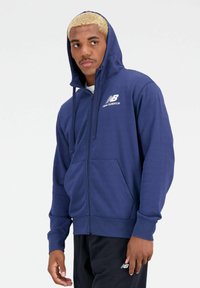 Marineblauwe zip-up hoodie van zachte stof, met een voorzak, trekkoord capuchon en wit New Balance-logo op de borst.