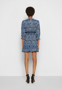 Mujer con peinado afro vestida con un vestido azul de estampado y mangas largas, de pie de espaldas, con zapatos de tacón alto negro con tachuelas.