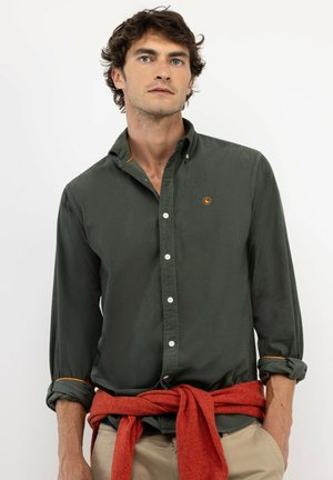 Hombre con camisa verde oscuro abotonada y suéter rojo atado alrededor de la cintura, de pie frente a un fondo liso.