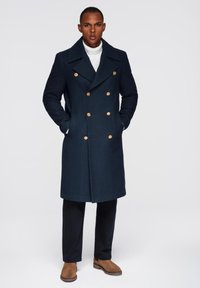 Marineblauer, doppelt geknöpfter Wollmantel mit goldenen Knöpfen, großem Kragen, Seitentaschen, getragen über einem weißen Rollkragenpullover und schwarzen Hosen, kombiniert mit braunen Schuhen.