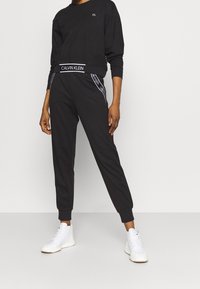 Sweat-shirt noir en coton à manches longues et col rond, associé à un pantalon de survêtement noir avec un logo "Calvin Klein" blanc sur la ceinture.