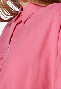 Camicia button-up rosa chiaro con colletto, caratterizzata da una texture liscia e una vestibilità morbida, con cuciture minime e un lieve lucido.