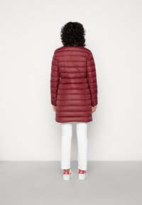 Abrigo acolchado granate con costuras horizontales, mangas largas y diseño de longitud media. Combinado con pantalones blancos y zapatos con detalles en rojo.