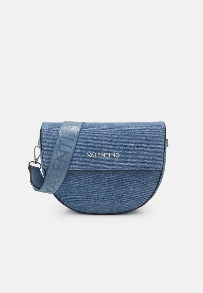 Denimová crossbody taška s kulatým tvarem. Má logo na přední straně, modrý popruh s vyrýtým designem a magnetické zavírání na klapce.