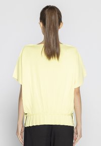 Jasnożółta plisowana bluzka z luźnymi krótkimi rękawami, dopasowaną talią i fakturowanym materiałem, widok z tyłu. Widoczne czarne spodnie.