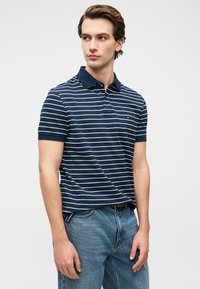 Μπλε ναυτικό ριγέ πόλο με κοντά μανίκια και γιακά. Ξεχωριστό κόκκινο λογότυπο στο στήθος. Φορεμένο με μπλε τζιν παντελόνι.