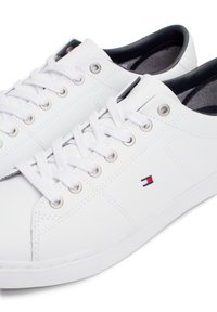 Baskets basses en cuir blanc avec lacets blancs et œillets en métal, avec un petit logo rouge, blanc et bleu sur le côté.