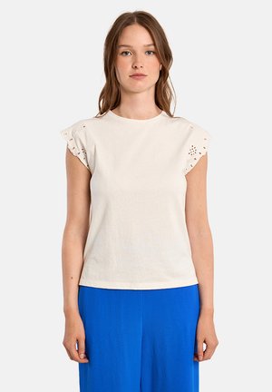 Donna con capelli castani lisci, indossa una maglietta bianca a maniche corte con dettagli a occhiello e pantaloni blu brillanti, in piedi contro uno sfondo semplice.