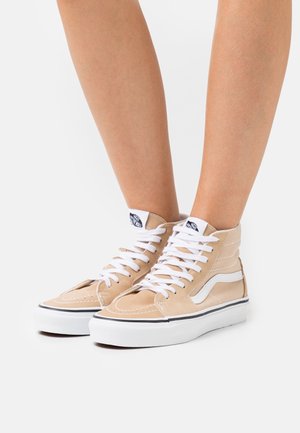 Baskets montantes beige avec lacets et semelles blancs portées aux pieds, arborant un logo Vans noir et blanc sur la languette.