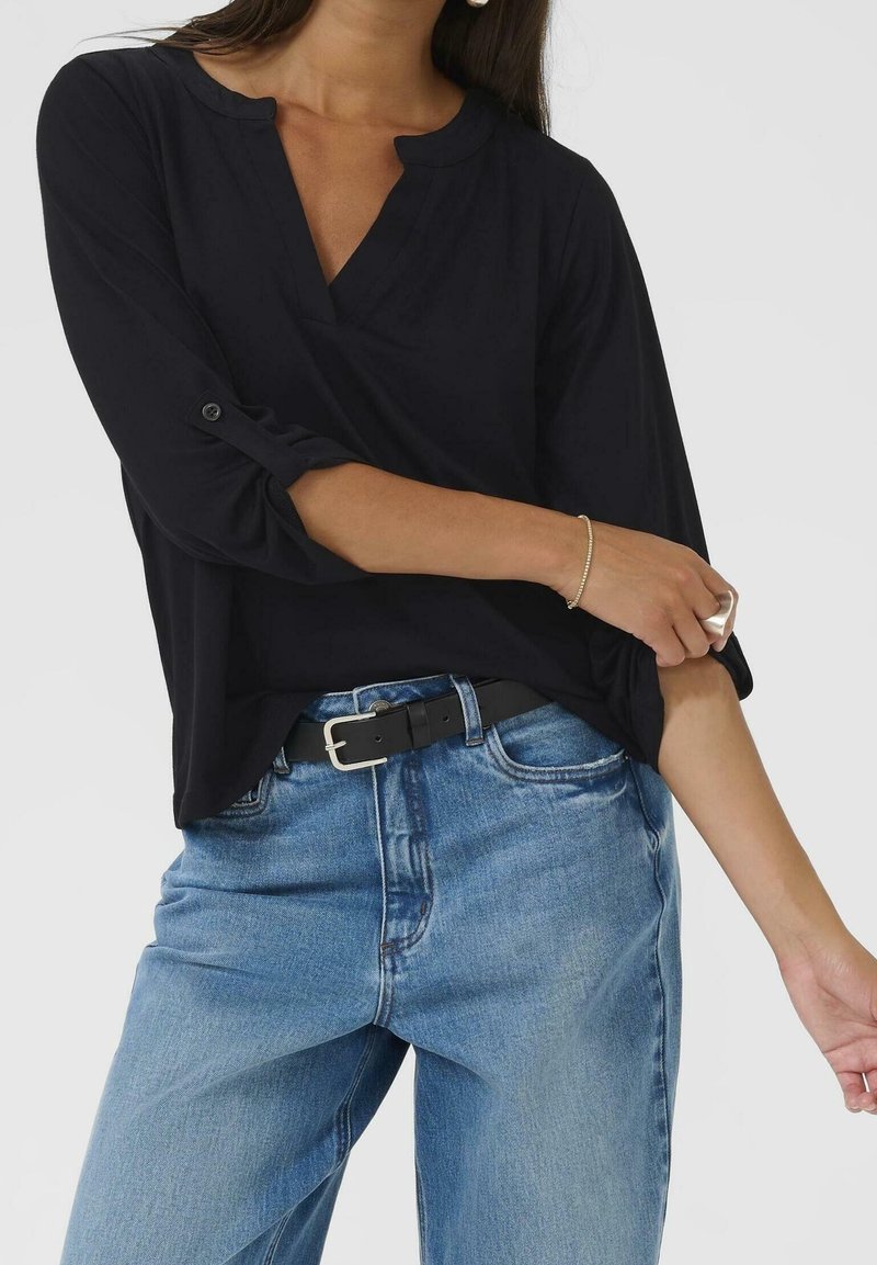 Femme portant un haut noir à manches longues retroussées et un jean bleu avec une ceinture noire, ajustant sa manche sur un fond blanc.