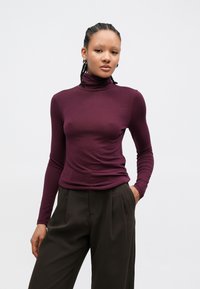 Μπλούζα με μακριά μανίκια και ζιβάγκο σε βαθύ μωβ χρώμα, φτιαγμένη από λείο και ελαστικό ύφασμα. Συνδυασμένη με παντελόνι ψηλόμεσο, άνετης εφαρμογής σε σκούρο καφέ χρώμα.