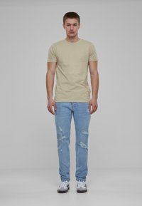 Camiseta de algodón beige, jeans azules desgastados, zonas usadas y decoloración, zapatillas blancas y grises. El modelo está de pie contra un fondo sencillo.