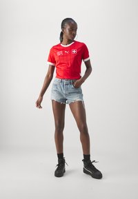 Sarkans īsām piedurknēm krekls ar baltiem akcentiem un Šveices karogu, apvienots ar nolietotām džinsa šortiem un melniem augstiem sporta apaviem.