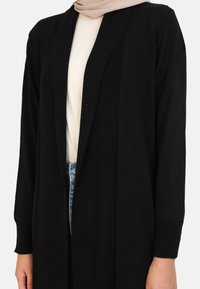 Zwarte, lange cardigan gemaakt van zachte stof, met een open voorkant en geribbelde mouwen. Draagt over een lichtgekleurde top en denim jeans.
