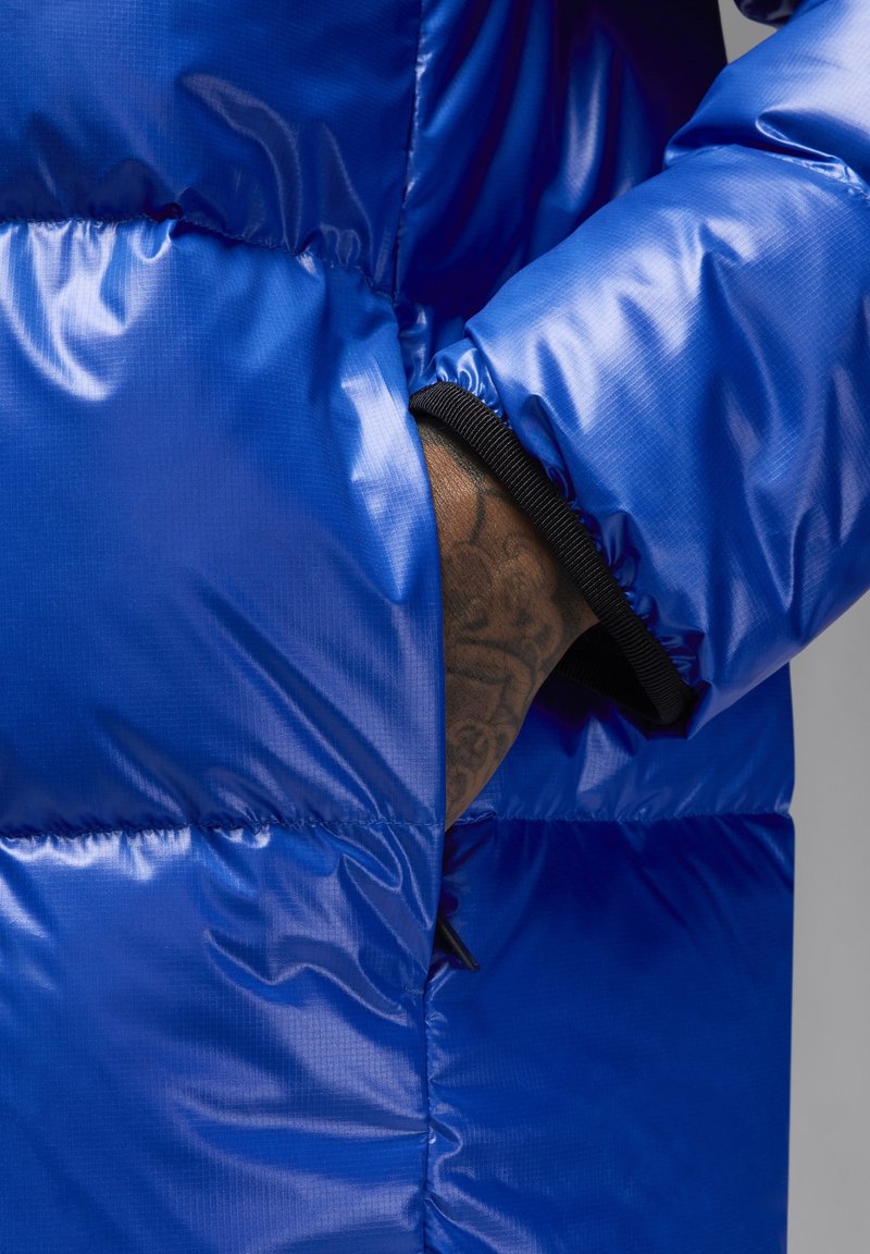 Chaqueta acolchada azul con una textura brillante y acolchada, con un bolsillo lateral y puños elásticos en negro contrastante para un ajuste y comodidad óptimos.