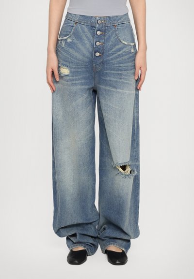 Jeans en denim bleu clair à jambe large avec une fermeture à plusieurs boutons, des zones déchirées et une découpe marquée au bas de la jambe.