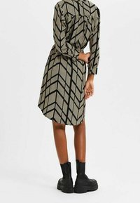 Femme portant une robe beige mi-longue à motif géométrique noir et des bottines noires à semelle épaisse, se tenant avec un bras plié derrière le dos.