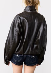 Giacca in pelle nera con collo alto e spalle scese, caratterizzata da una texture liscia e una vestibilità rilassata, abbinata a pantaloncini di jeans blu.