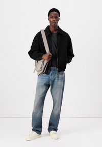 Black denim jacket, black shirt, light blue jeans, and white sneakers. Gray shoulder bag with strap, standing against a plain background.