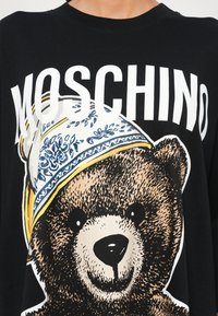 T-shirt noir avec un grand ours en peluche portant une écharpe bleue et jaune à motifs, avec le texte "MOSCHINO" au-dessus de l'ours.