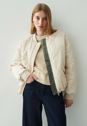 Beige genre-quilted bomberjack met groene accenten, geribbelde manchetten en een ritsluiting, gedragen over een lichtbeige trui en donkerblauwe wijde broek.