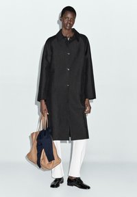 Cappotto nero oversize con chiusura a bottoni, tasche laterali e colletto a risvolto; abbinato a pantaloni bianchi e scarpe nere; porta borse beige e blu navy.