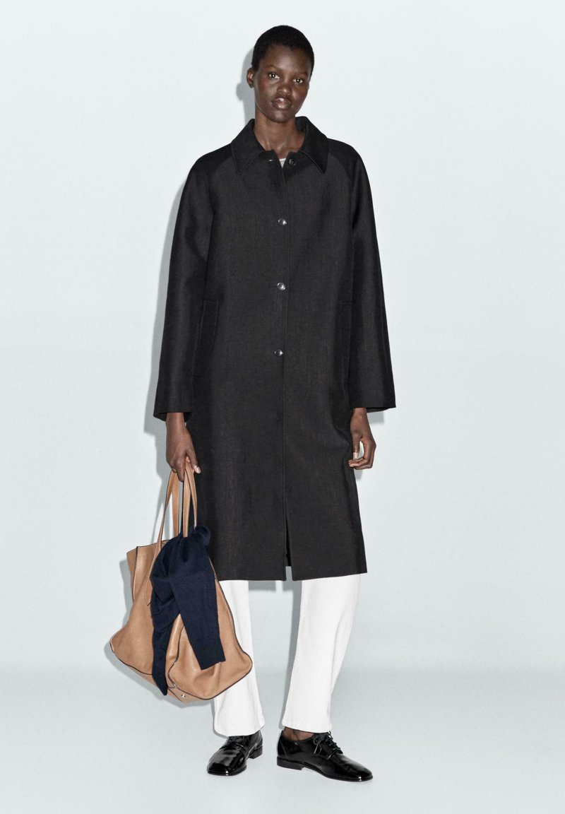 Cappotto nero oversize con chiusura a bottoni, tasche laterali e colletto a risvolto; abbinato a pantaloni bianchi e scarpe nere; porta borse beige e blu navy.