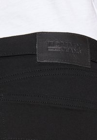 Jeans de mezclilla negra con una etiqueta de cuero con "TOMMY JEANS" grabada. Incluye detalles de costura visibles y una textura suave.
