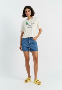Witte korte mouwen shirt met kleurrijke rand, voorzien van een geborduurd cartoonfiguur en cactussen ontwerp, gecombineerd met blauwe denimshorts en gele sliders.