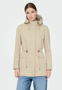Veste imperméable beige avec capuche, cordons de serrage ajustables à la taille et deux poches avant. Texture lisse et design décontracté.