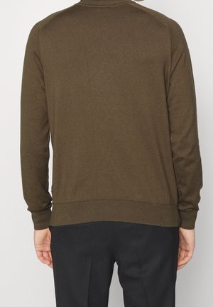 Ruskea neule, jossa on ribatut kaulus ja hihansuut, sileällä pinnalla ja vartalonmyötäisellä muotoilulla, näkyy takaa yksinkertaista taustaa vasten.