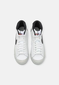 Höga sneakers i vitt läder med svarta detaljer, som har en grå tåhuvud, vita snören och en röd logotyp på tungan.