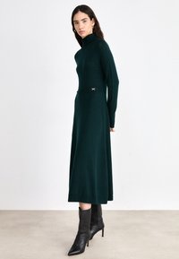 Strickkleid in Dunkelgrün mit hohem Kragen, langen Ärmeln und taillierter Taille. Verfügt über goldene Akzentdetails an der Taille und einen ausgestellten Rock.