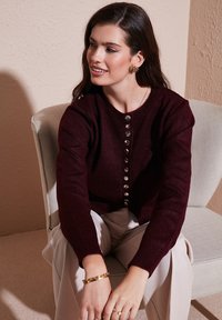 Gilet en tricot bordeaux avec un col rond, des manches longues et des boutons métalliques sur le devant, accompagné de pantalons clairs.