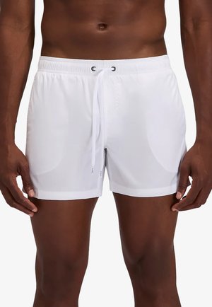 Busto maschile che indossa pantaloncini da bagno bianchi con coulisse in vita, in piedi davanti a uno sfondo semplice.