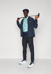 Veste coupe-vent marine sur un polo à motifs bleu clair, pantalon sombre et chaussures blanches. Tenant un club de golf, portant une casquette marine assortie.