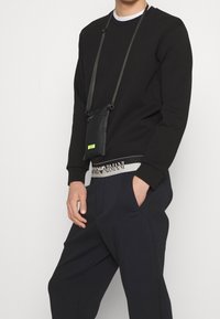 Czarny sweter z okrągłym dekoltem i prążkowanymi mankietami, w zestawie z granatowymi spodniami. Mała czarna torebka na biodro z neonowym logo wisi na szyi.