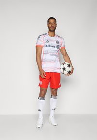 Futballista fehér és rózsaszín mintás mezben, piros nadrágban, fehér zokniban fekete csíkokkal, és egy fekete-fehér focilabdát tart a kezében.