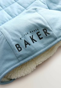 Jasnoniebieska pikowana tkanina z miękkim białym futerkiem. Czarny napis "BY TED BAKER BAKER" wskazuje na markę.