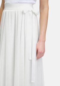 Jupe plissée blanche avec une taille lisse et un détail en nœud. Fabriquée dans un tissu léger avec une texture douce et transparente.