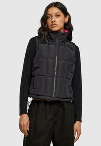 Svart puffer vest med høy krage, glidelås og kviltet design. Brukes over en langermet svart topp med slank passform og cropped lengde.