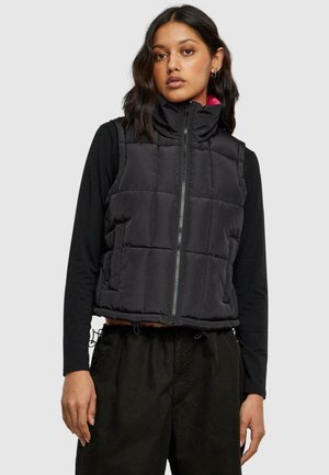 Svart puffer vest med høy krage, glidelås og kviltet design. Brukes over en langermet svart topp med slank passform og cropped lengde.