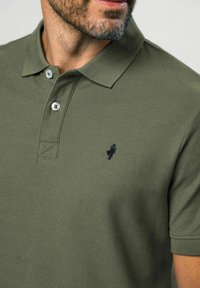 Tricou polo verde măsliniu din bumbac, cu guler, două nasturi și un logo brodat albastru închis pe pieptul stâng. Material texturat.