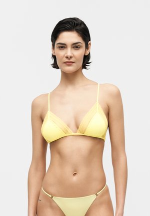 Rövid sötét hajú nő sárga csipkés bikinifelsőt és hozzá illő bikinialsót visel egyszínű háttér előtt.