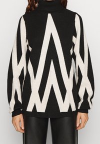 Schwarzer und weißer Rollkragenpullover mit auffälligen Zickzackmustern, gestricktem Textur und übergroßen Ärmeln, kombiniert mit schwarzen Lederhosen.